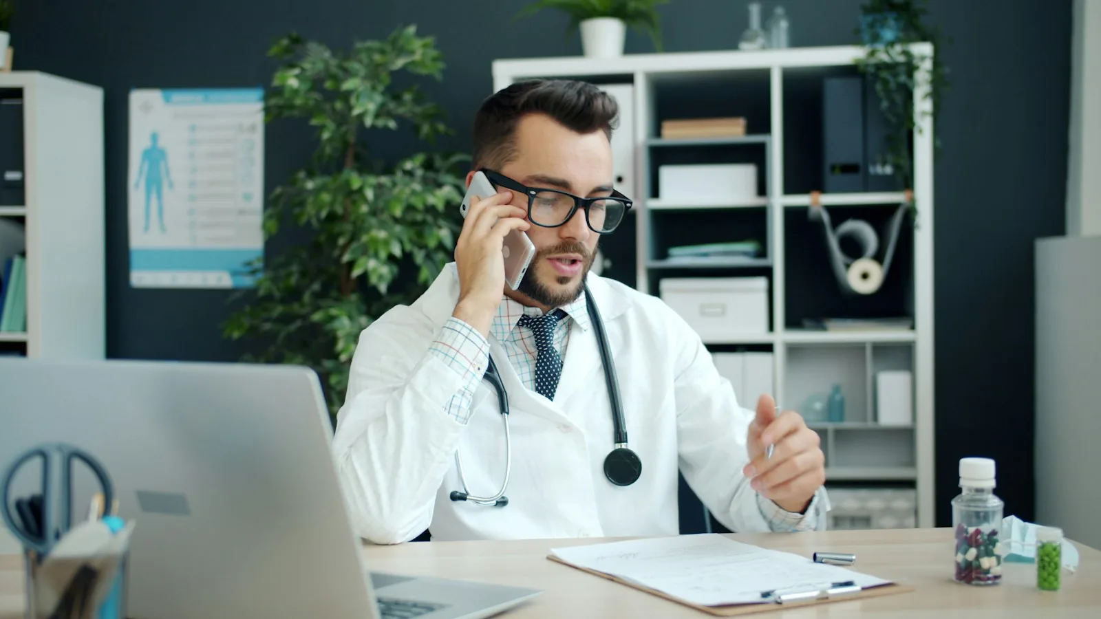 Doctor in a white coat taking a phone call at his office desk beside a laptop