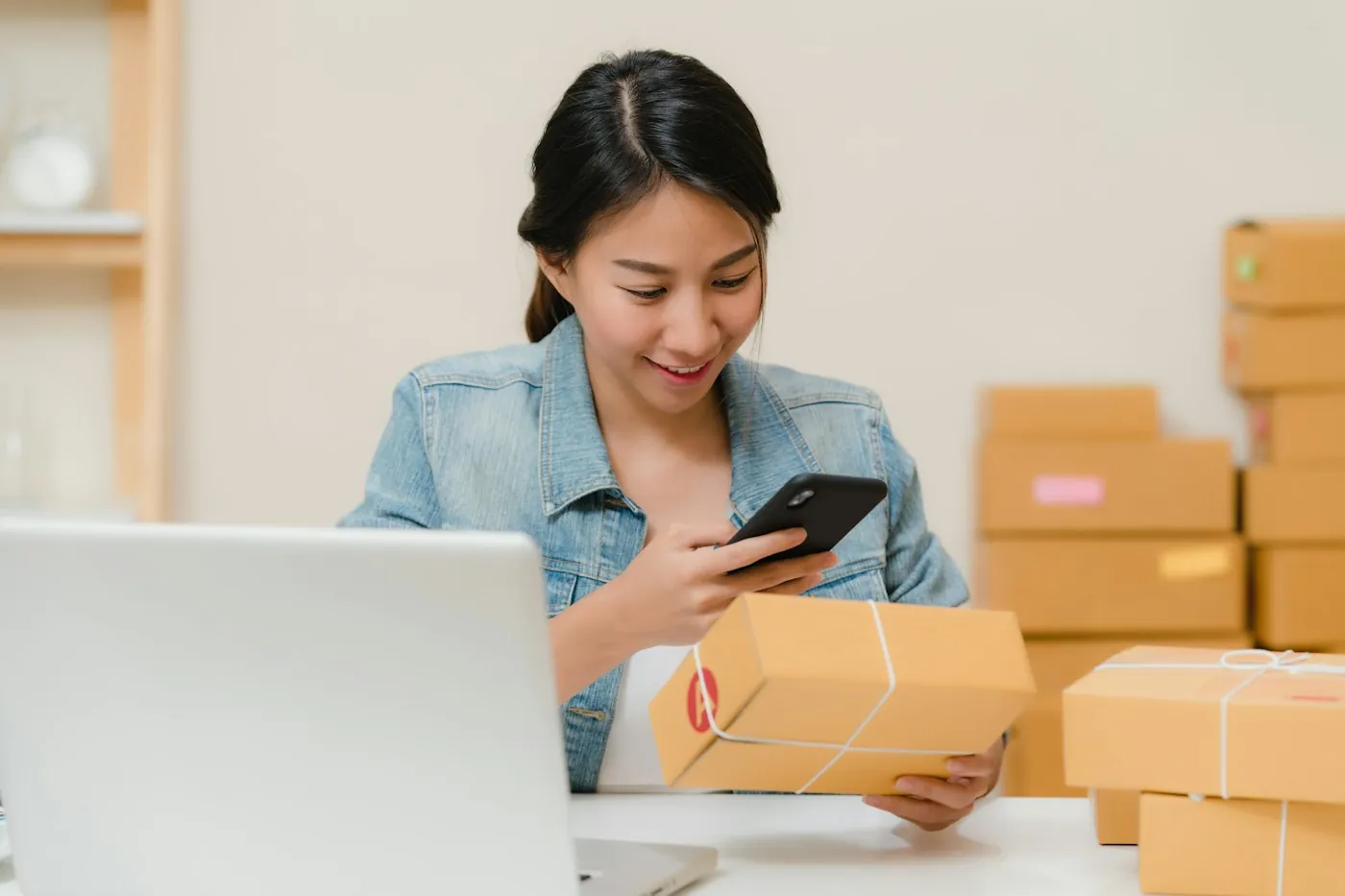 Online store owner checking orders on a phone beside parcels and a laptop